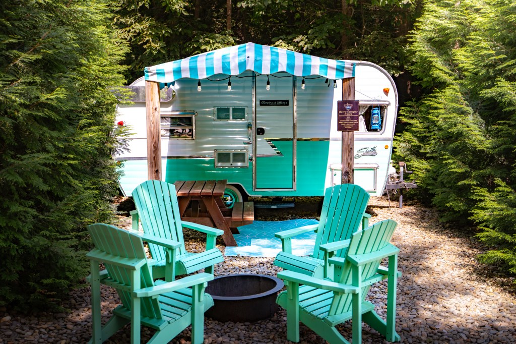 A vintage teal and white camper surrounded by lush greenery, featuring a striped awning and outdoor seating area with wooden chairs and a fire pit.