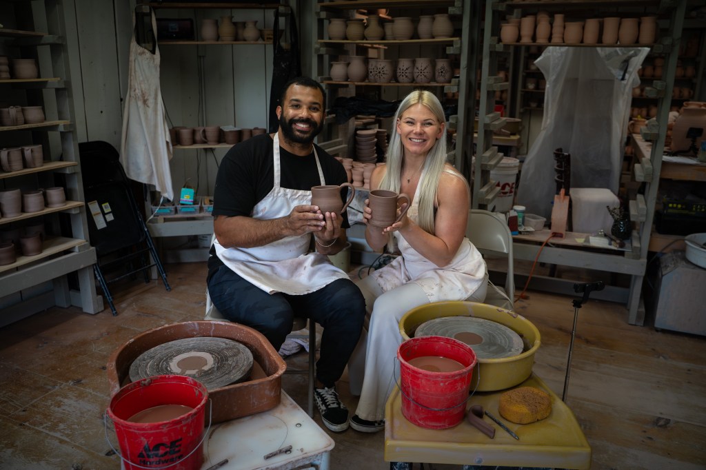 A smiling man and woman sitting in a pottery studio, each holding a freshly made clay mug. The studio background features shelves filled with various ceramic pieces.