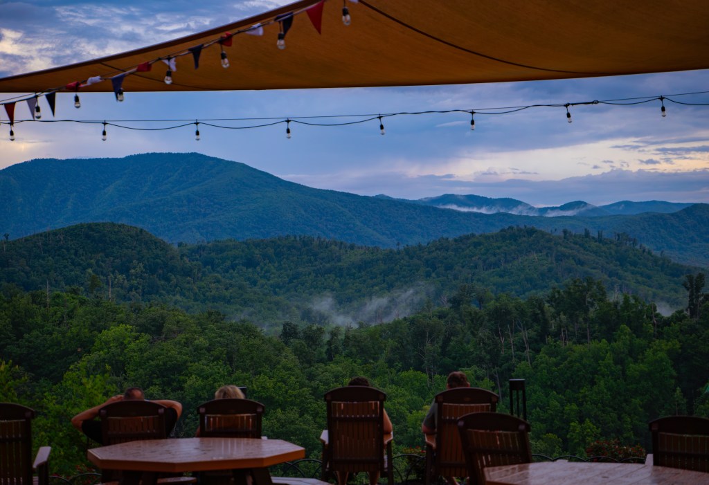 View of rolling green mountains under a cloudy sky, with string lights and a canopy in the foreground, and people seated at wooden tables.