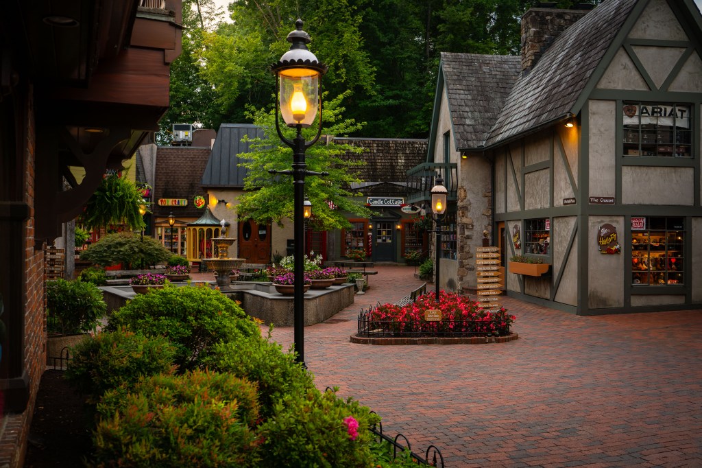 A charming shopping village with cobblestone pathways, colorful storefronts, and flower beds. Street lamps cast a warm glow, and a variety of shops can be seen, including a toy store and a candle shop.