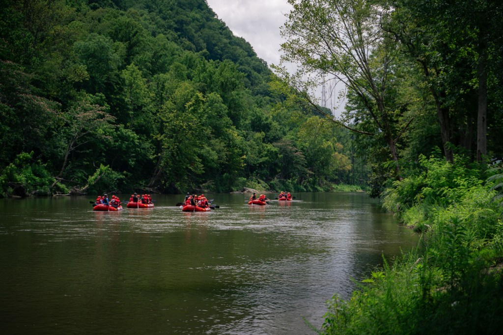 A group of people in red rafts paddling down a calm river surrounded by lush green trees and hills.