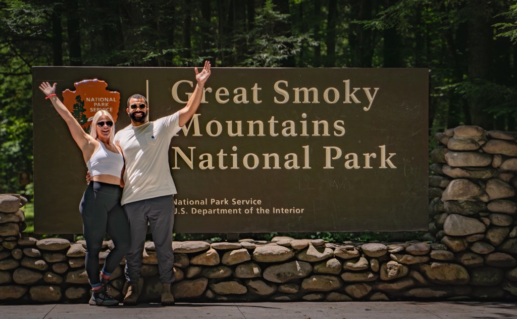 A man and a woman pose joyfully in front of the Great Smoky Mountains National Park sign, surrounded by lush trees.