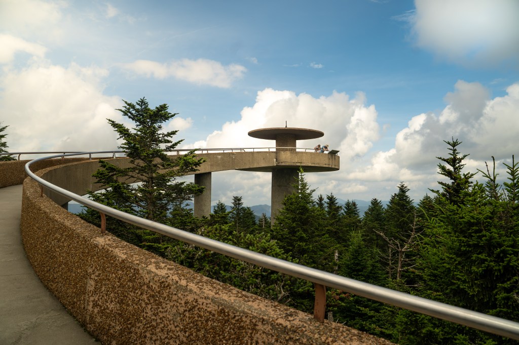 A scenic overlook with a circular viewing platform atop a concrete structure, surrounded by tall evergreen trees and a blue sky with clouds.